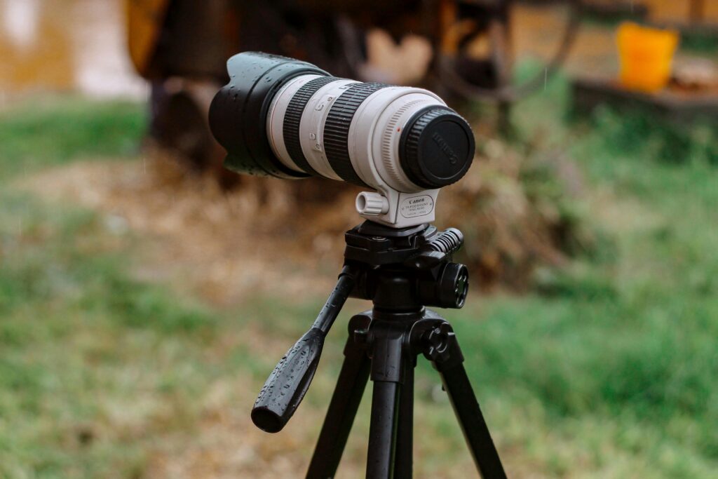Camera lens mounted on a tripod outdoors, raindrops visible on the lens and body of the setup.
