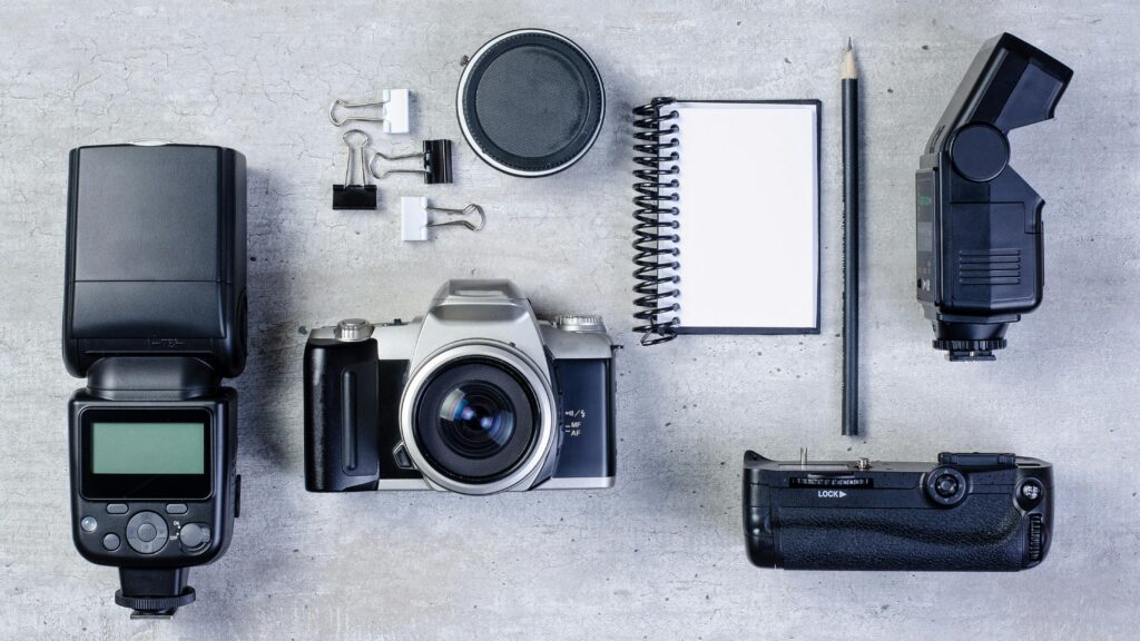 Flat lay of camera gear: two cameras, a lens, a flash unit, binder clips, a spiral notebook, and a pencil on a gray textured surface.