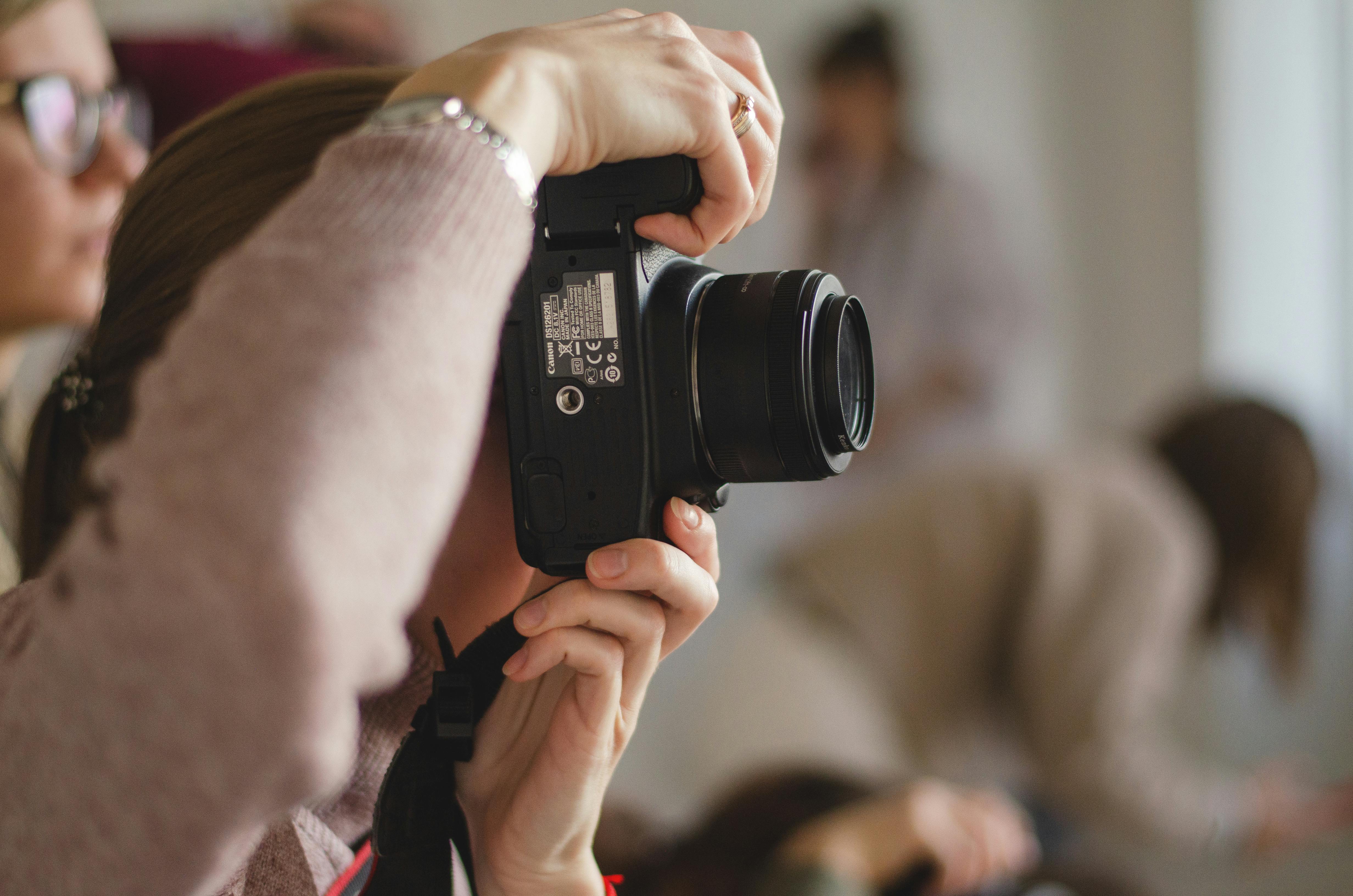 Close-up of a photographer holding a DSLR camera to their eye, ready to shoot at an event or gathering in a softly blurred background.