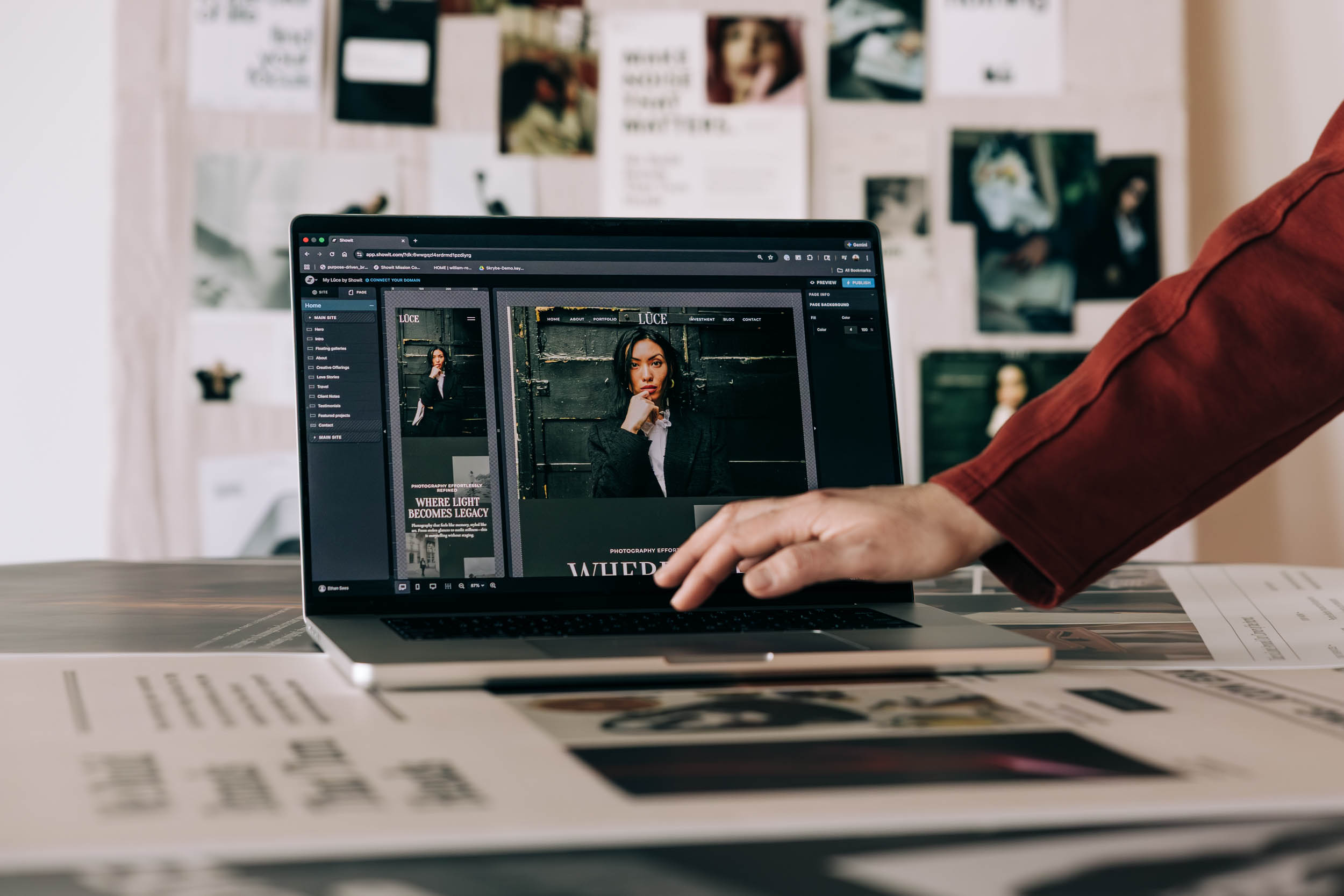 Person in a red sleeve uses a laptop to design a fashion magazine layout at a cluttered desk.