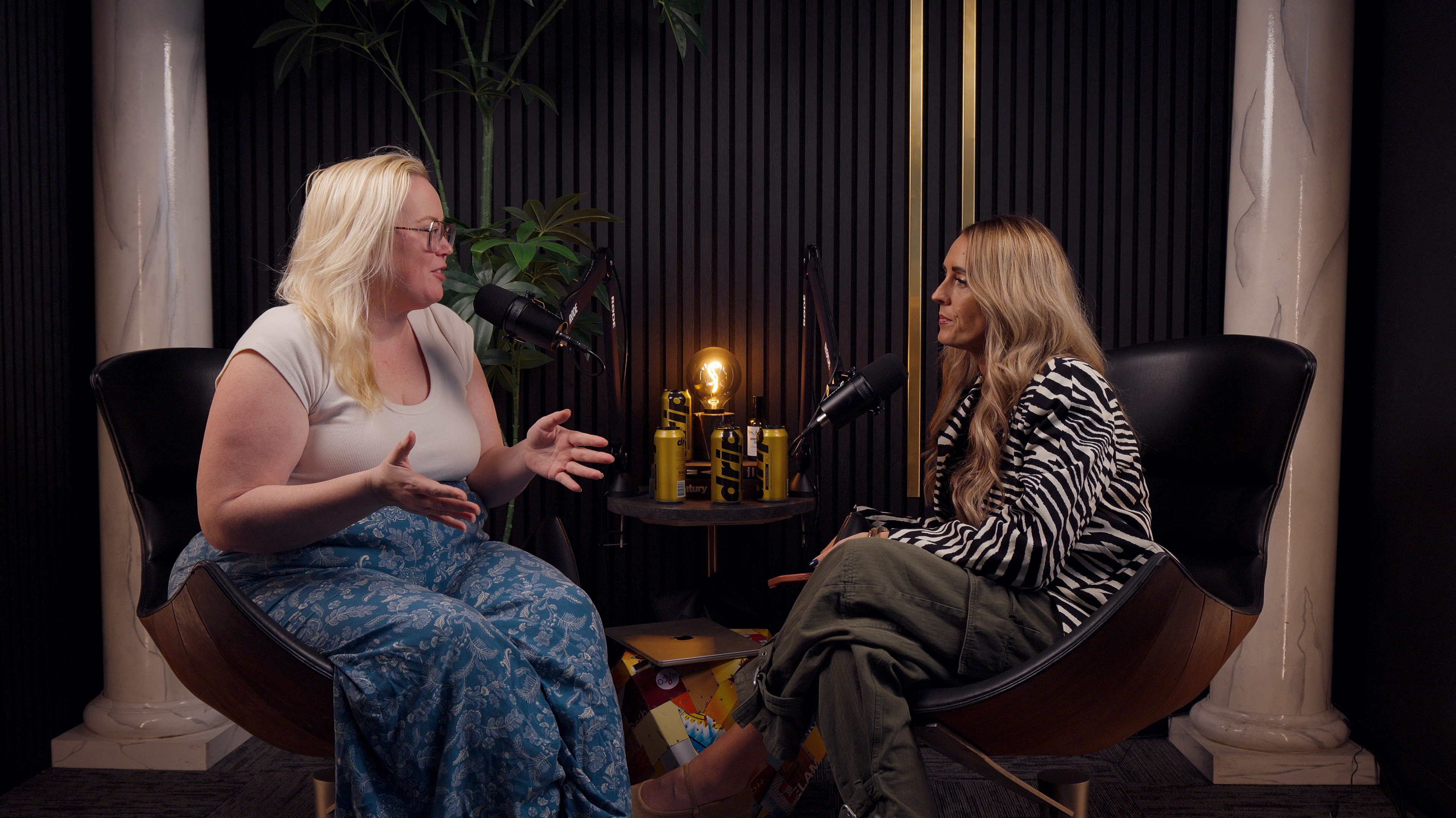 Two women sit in a podcast studio facing each other, speaking into microphones with drinks on the table between them.