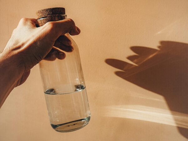 Hand gripping a glass bottle with a cork stopper filled with water, against a warm beige wall with a plant shadow to the right.