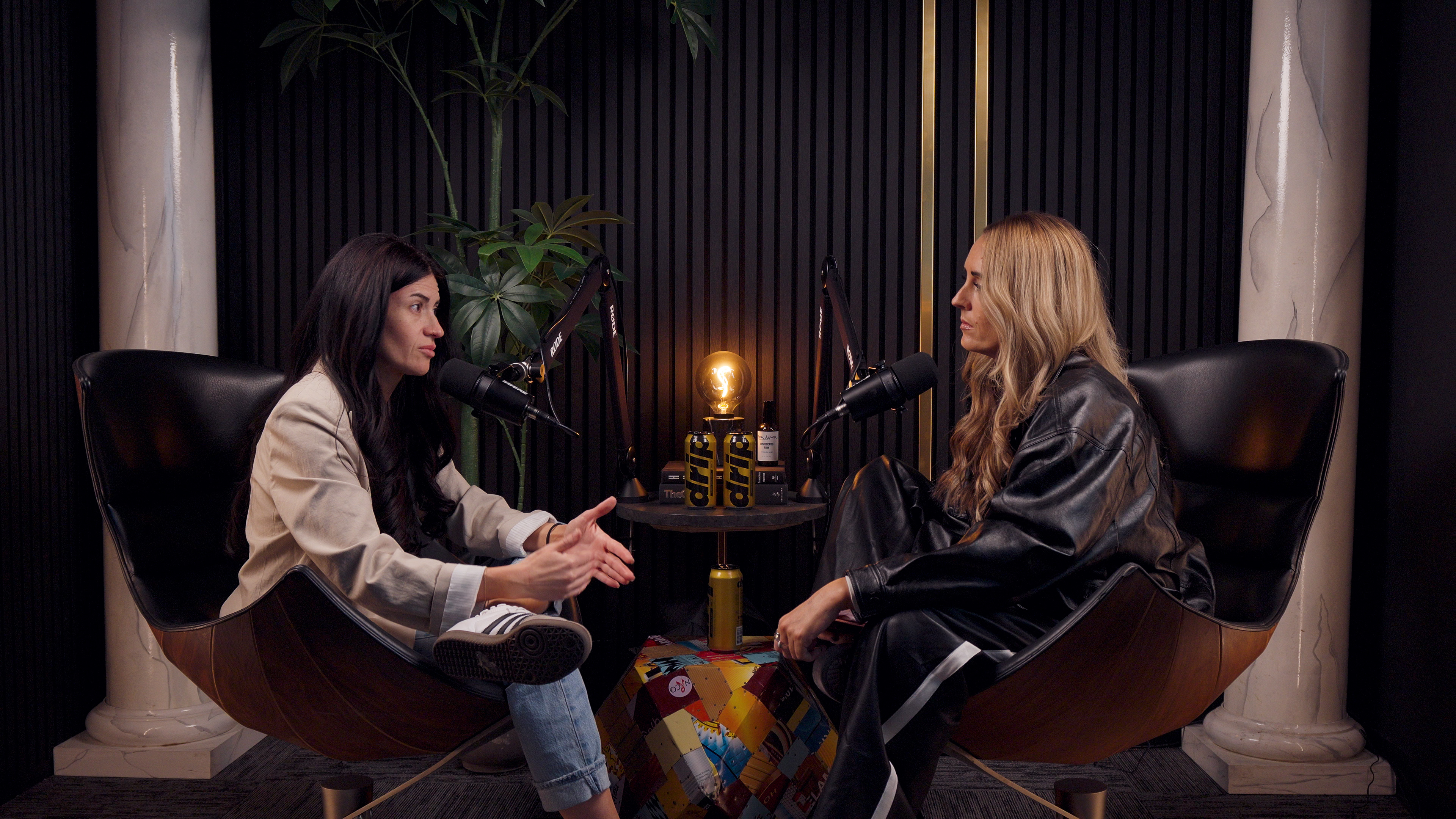 Two women are seated in curved chairs facing each other, speaking into microphones in a podcast studio.