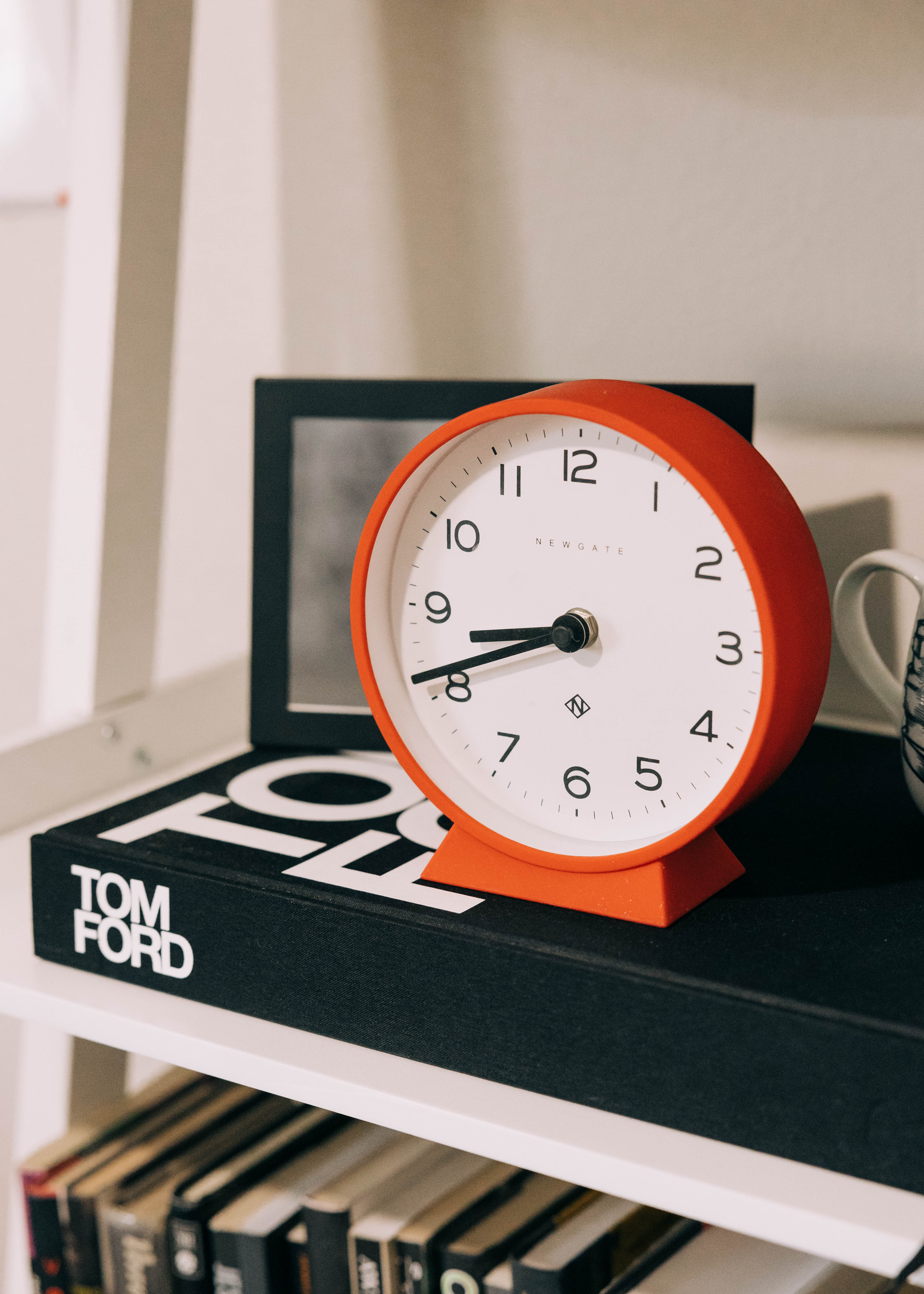 Red circular desk clock perched on a black Tom Ford box on a bookshelf shelf.