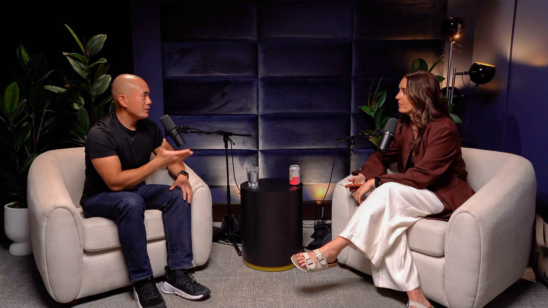 Two people sit in beige armchairs facing professional microphones in a podcast studio, with plants and a small table between them.