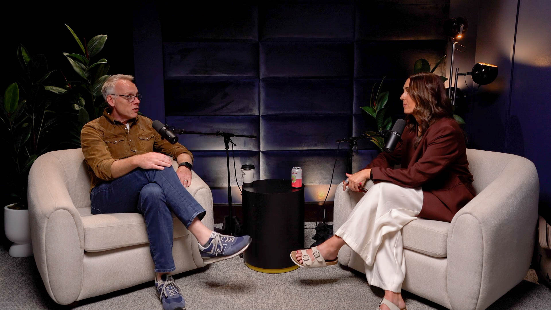 Two people sit on beige armchairs facing each other in a podcast studio, speaking into handheld mics with a small table and beverages between them.