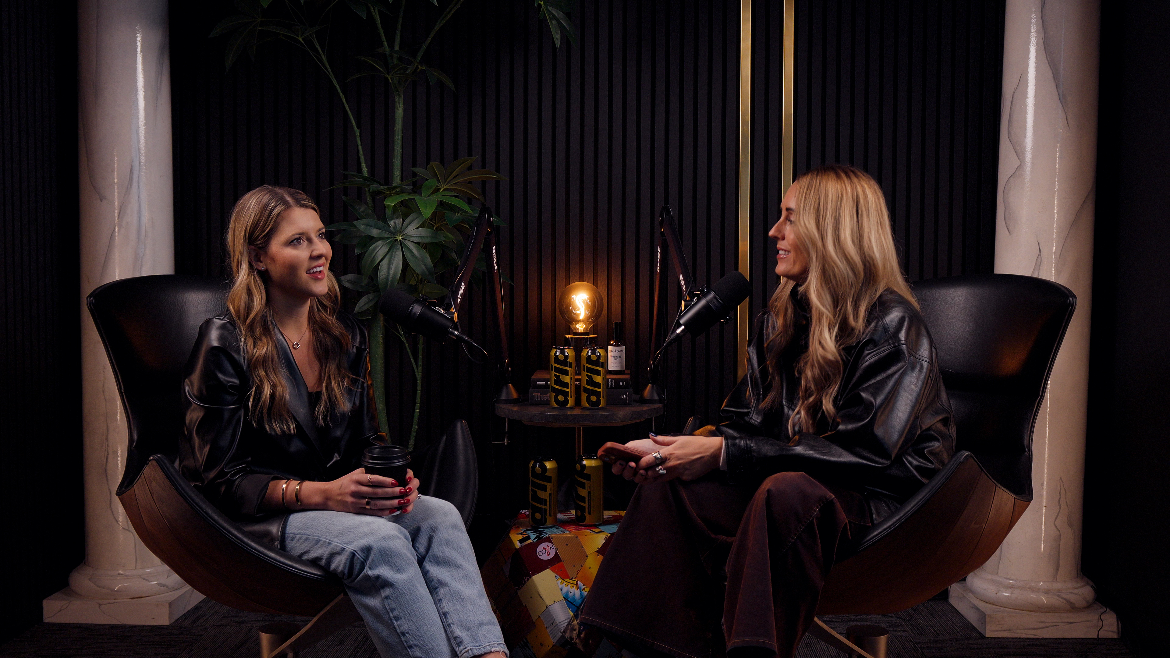 Two women wearing black jackets sit in modern chairs across from each other in a podcast studio, speaking into microphones with a warm light between them.