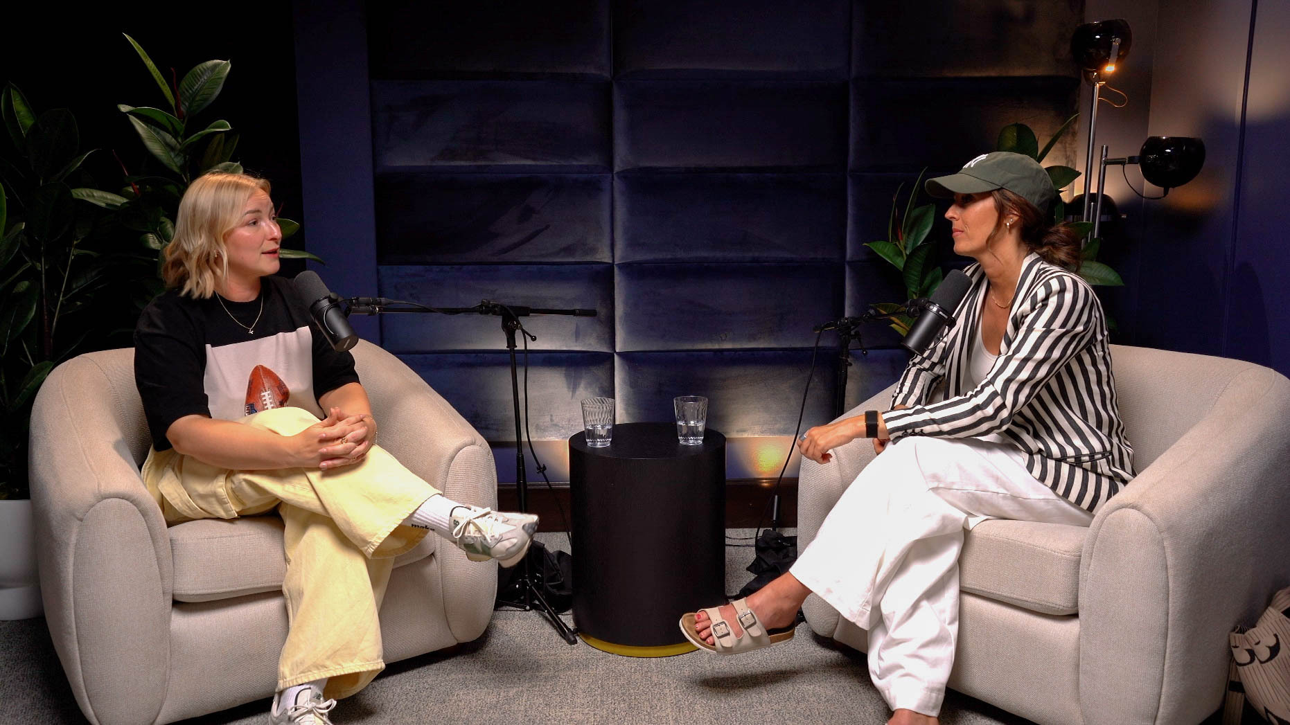 Two women are interviewed in a podcast studio, seated in beige chairs with microphones between them.