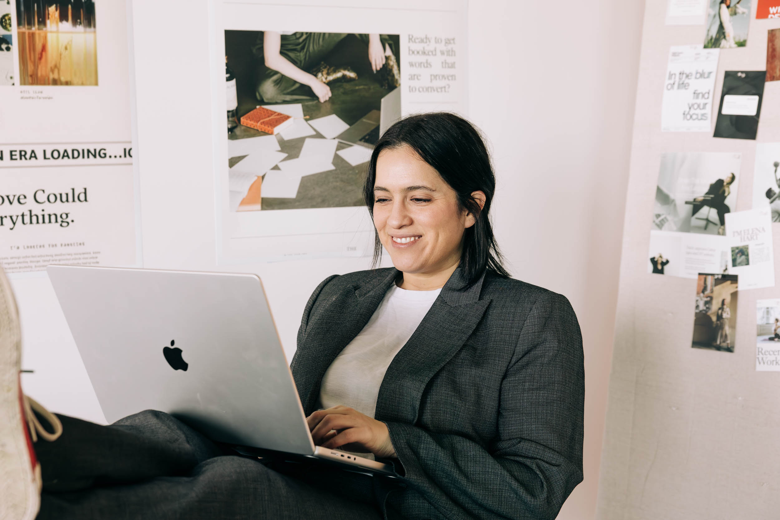Smiling business owner in a gray blazer typing a blog post on her laptop, with printed website page designs pinned to the wall