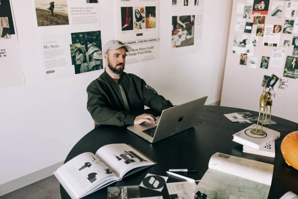 Creative business owner in a ball cap focused on his laptop at a desk covered in open design books and magazines reading who is showit for