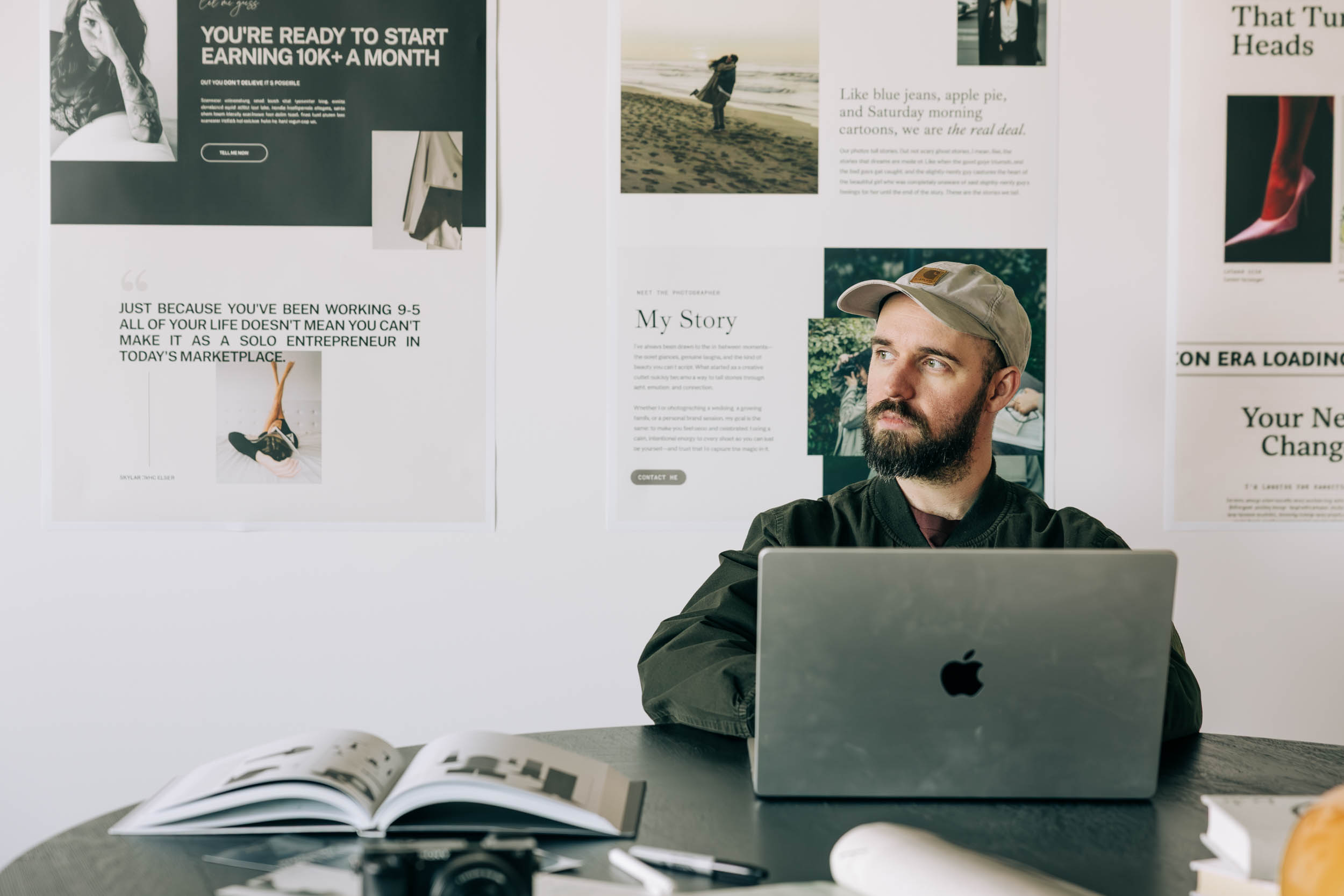 Bearded creative business owner in a ball cap at his laptop, with a camera and photo book on the desk and website designs pinned behind him