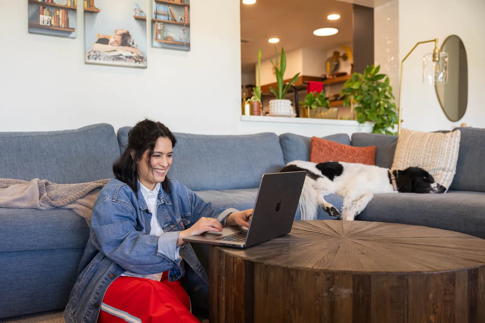 Woman smiling while working on a laptop at a coffee table in a living room, with a dog asleep on the couch behind her
