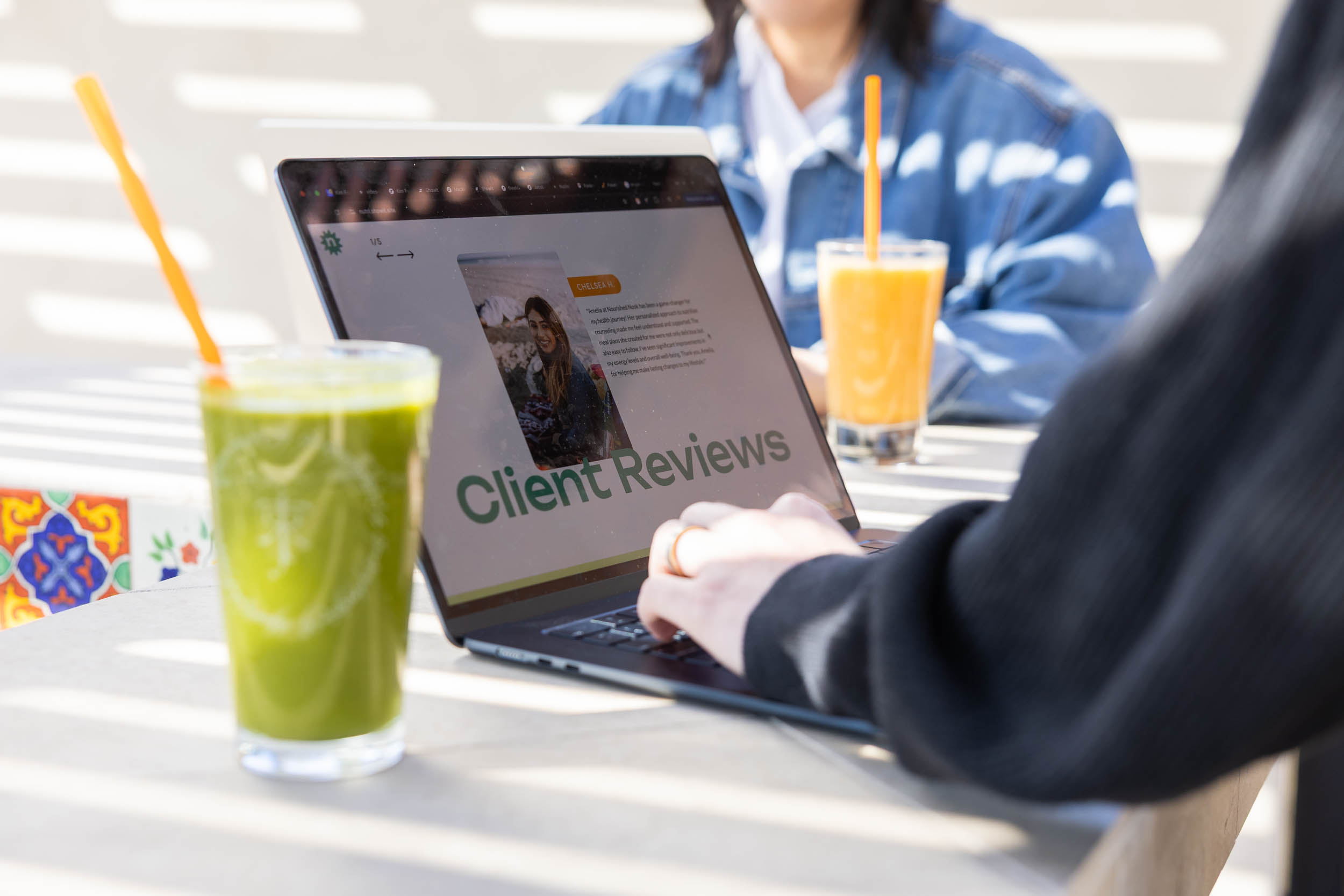 Person working on the client reviews section of a wellness website on a laptop at a sunny outdoor table with fresh juices