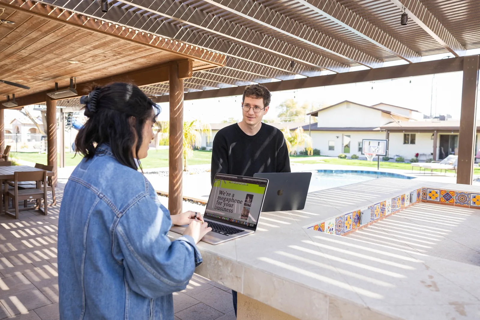 Two people talk at an outdoor patio counter while one works on a website on a laptop