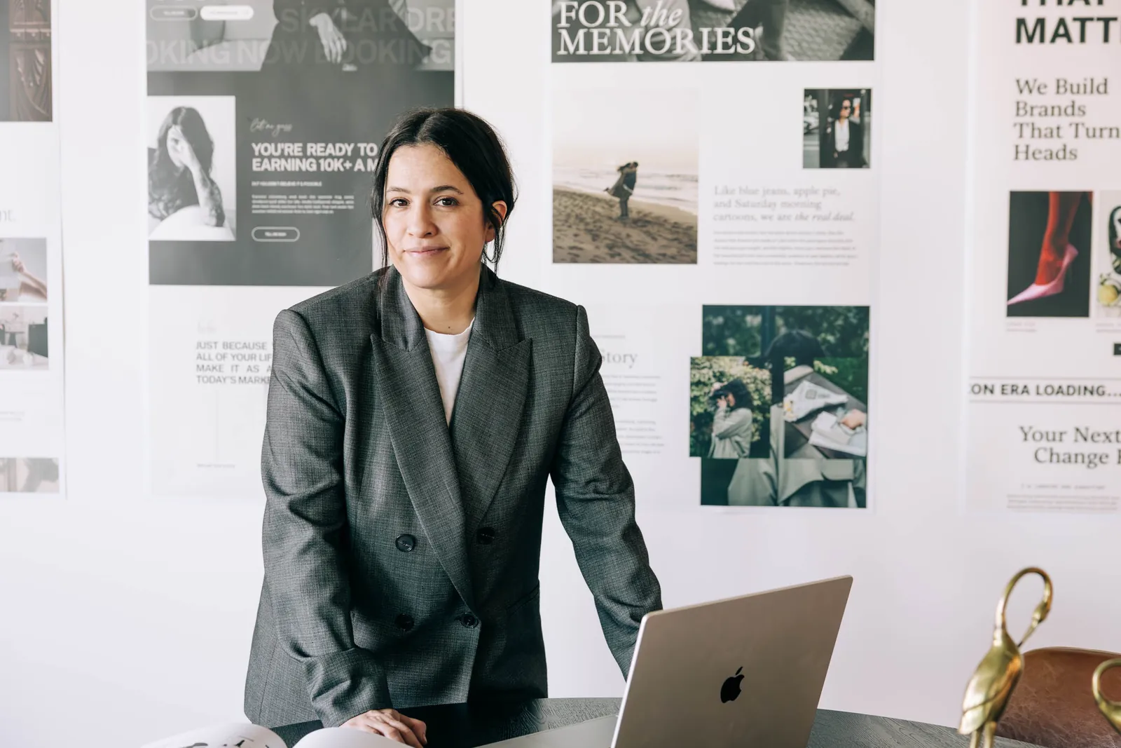 website-design-wall-laptop-referrals - Showit Woman in a gray blazer stands at a desk with a laptop in front of a wall of printed website page designs